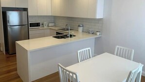 A modern kitchen with white cabinets, a stainless steel refrigerator, a microwave, and a sink. There is a white dining table with chairs.