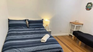 A neatly made bed with striped bedding, a bedside lamp, a small wooden nightstand, and a bench with a black cover.
