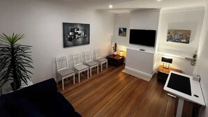 A modern living room with a wooden floor, white walls, and a flat-screen TV mounted on the wall. There are several white chairs arranged in a row, a small table with a lamp, and a potted plant.