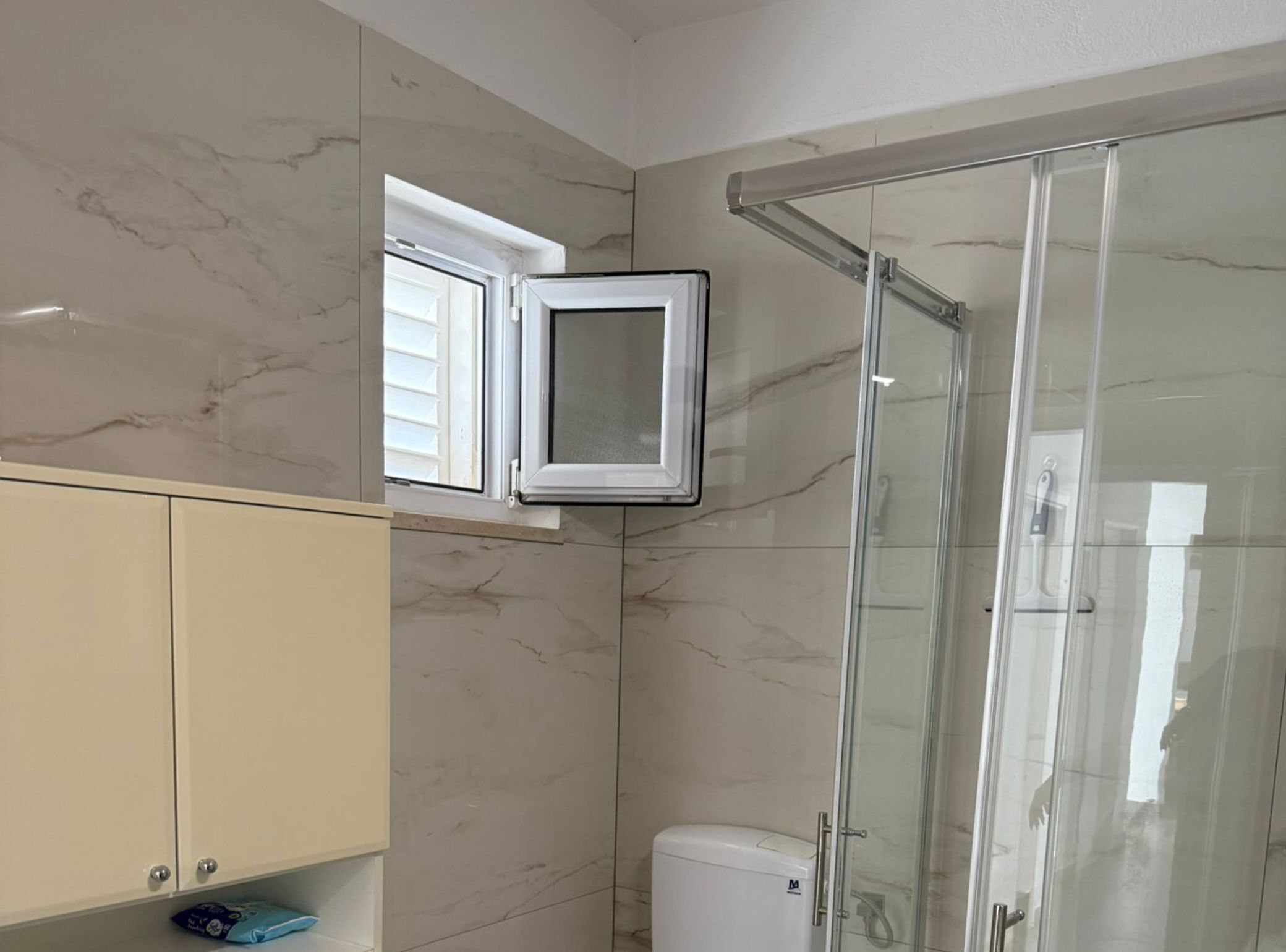 Un bagno con box doccia in vetro, un water e un mobile con una finestra sopra.