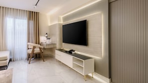 Une chambre d’hôtel moderne avec un téléviseur à écran plat fixé au mur, une console blanche, un fauteuil beige et une grande fenêtre avec des rideaux.