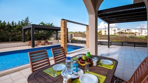 An outdoor dining area with a pool, wooden chairs, and a table set for two with plates, glasses, and a fruit bowl. - Villa Blue Wave Tessera (Rhodes)