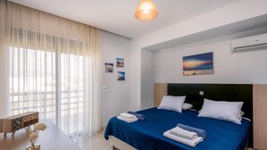 A modern hotel room with a large bed, a desk, and a view of the city through sheer curtains. - Villa Blue Wave Tessera (Rhodes)
