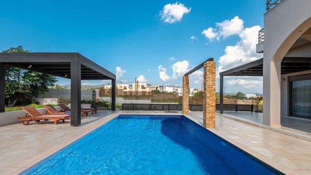 A swimming pool with lounge chairs, a stone pillar, and a view of buildings in the background. - Villa Blue Wave Tessera (Rhodes)
