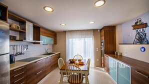A modern kitchen with wooden cabinets, a dining table with chairs, and a large window with sheer curtains. - Villa Blue Wave Tria (Rhodes)