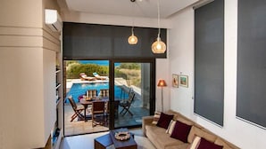 A modern living room with a large sofa, a coffee table, and a view of an outdoor pool area. - Villa Blue Wave Tria (Rhodes)