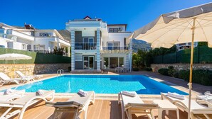 A swimming pool with lounge chairs and umbrellas, surrounded by a stone wall and greenery. - Four Seasons Villa in Oludeniz (Fethiye)