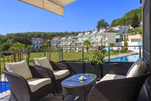 A balcony with wicker furniture, a glass table, and a view of a swimming pool and buildings in the background. - Residence Villa U in Oludeniz (Fethiye)