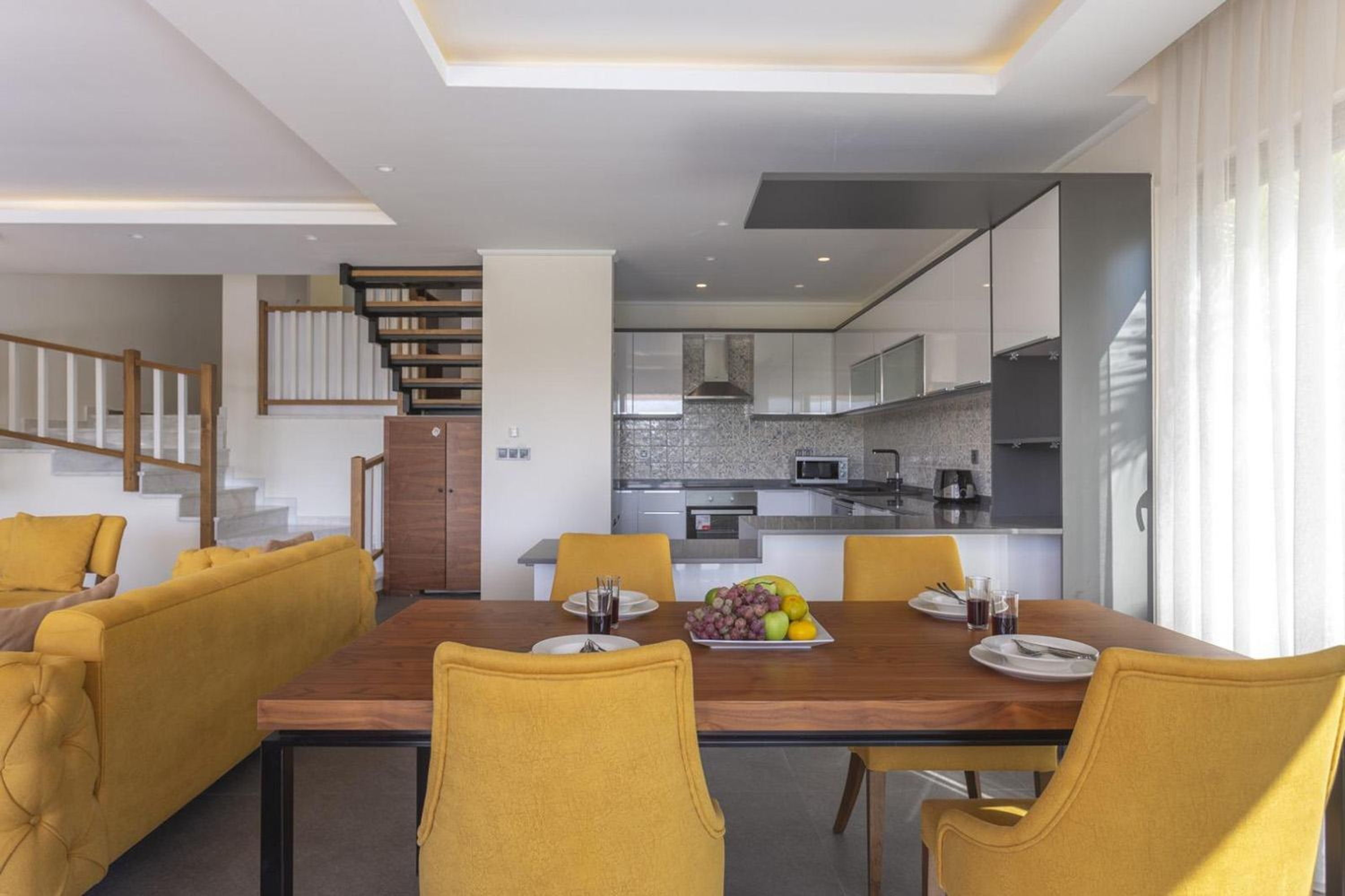 A modern kitchen with a central island, a dining area with yellow chairs, and a staircase leading to an upper level.