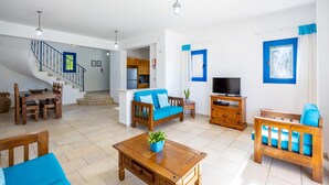 A spacious living area with a wooden sofa, a coffee table, and a TV stand. A staircase leads to an upper floor. - Villa Apollonas in Peyia (Pegeia)