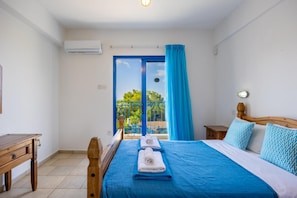 A bedroom with a bed, a wooden nightstand, a desk, and a window with a view of greenery. - Villa Apollonas in Peyia (Pegeia)