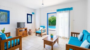 A spacious living room with a TV, wooden furniture, and a view of a pool through the glass door. - Villa Apollonas in Peyia (Pegeia)