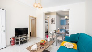 A modern living room with a TV, a wooden coffee table with fruits, a teal sofa, and a dining area with a glass table and chairs. - Villa Memi in Spartia (Kefalonia)