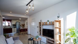 Una sala moderna con un sofá, un televisor y un mueble de entretenimiento de madera. En el fondo, se aprecia un comedor con una mesa y sillas.