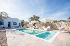En pool med solstolar, en stenmur och en vit byggnad med blå detaljer.