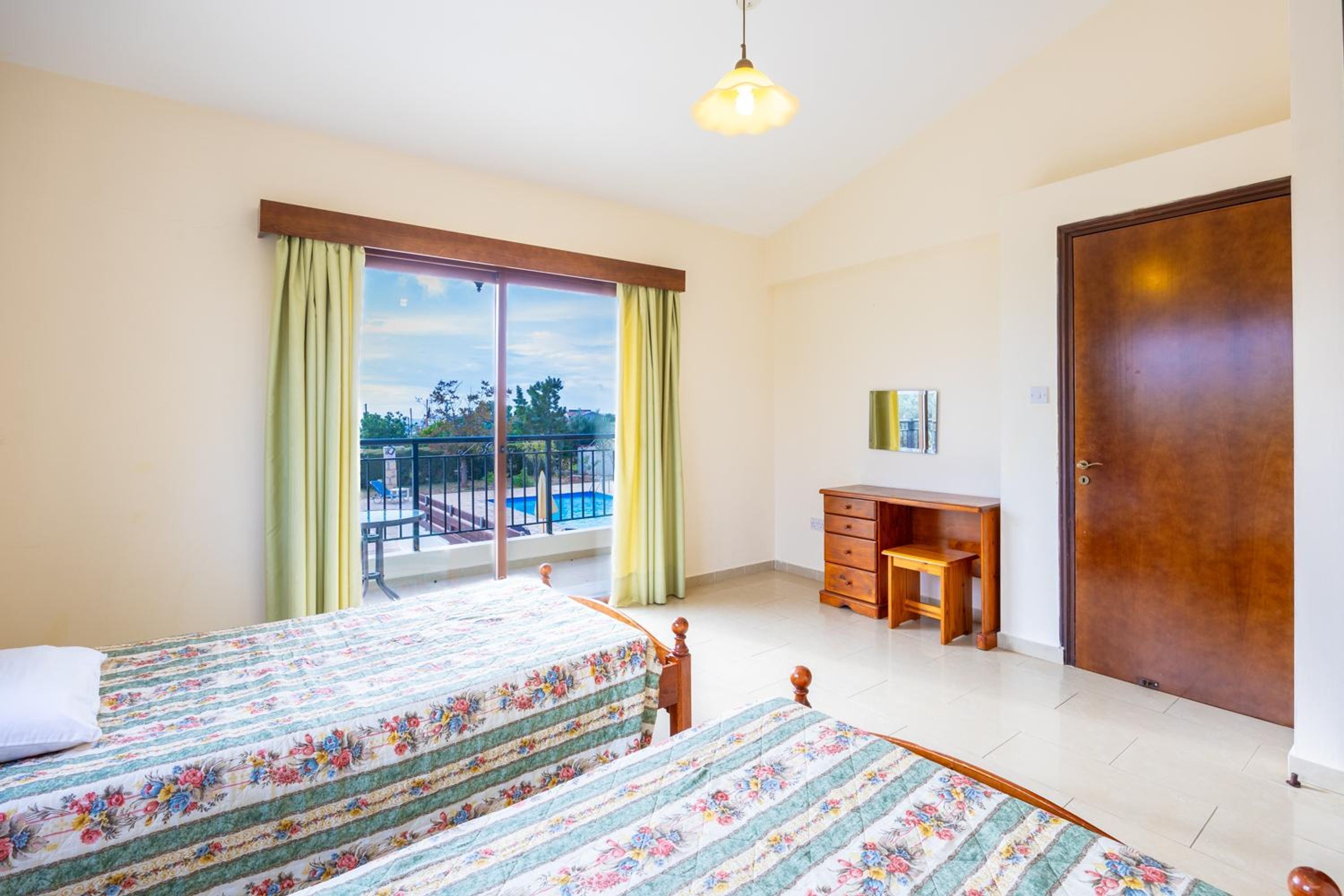 A bedroom with two beds, a desk, a chair, a wardrobe, and a balcony with a view of the outdoors.