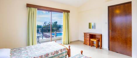 A bedroom with two beds, a desk, a chair, a wardrobe, and a balcony with a view of the outdoors.
