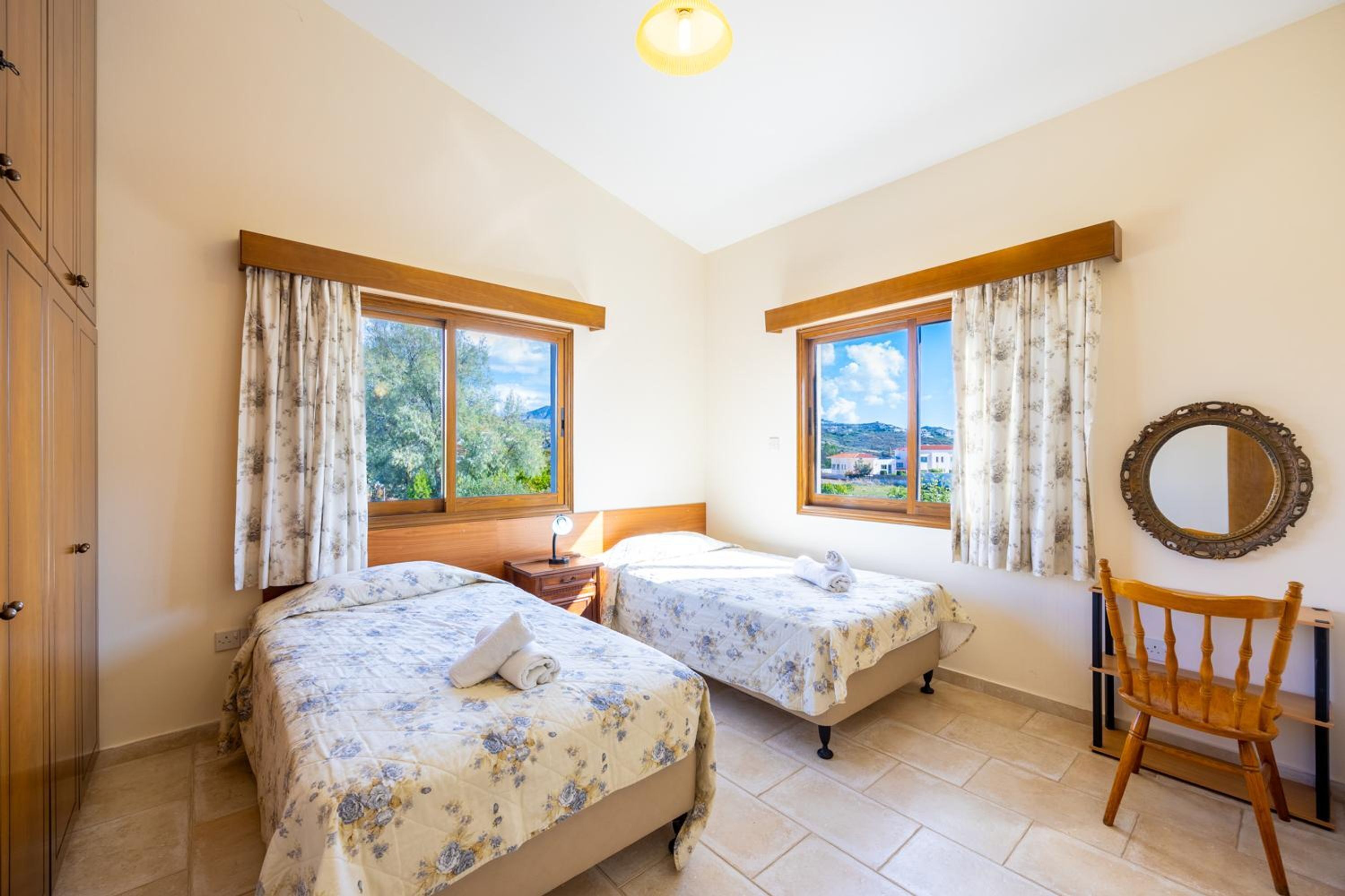 A bedroom with two beds, a wooden chair, a mirror, and large windows with curtains.