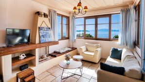 Ein gemütliches Wohnzimmer mit Fernseher, einem Holzregal, einer Couch, einem kleinen Tisch und Blick auf das Meer.