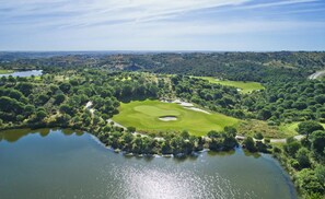 Un campo de golf con un estanque, rodeado de árboles densos y un paisaje ondulado.