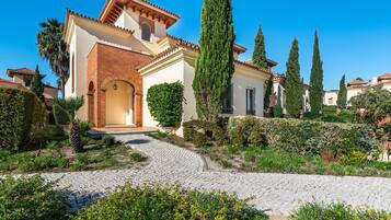 Uma villa tradicional em estilo mediterrâneo, com um caminho de pedras que leva à entrada, rodeada por sebes bem cuidadas e altas árvores de cipreste.