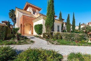 Una villa tradicional de estilo mediterráneo con un camino empedrado que lleva a la entrada, rodeada de setos cuidados y altos cipreses.