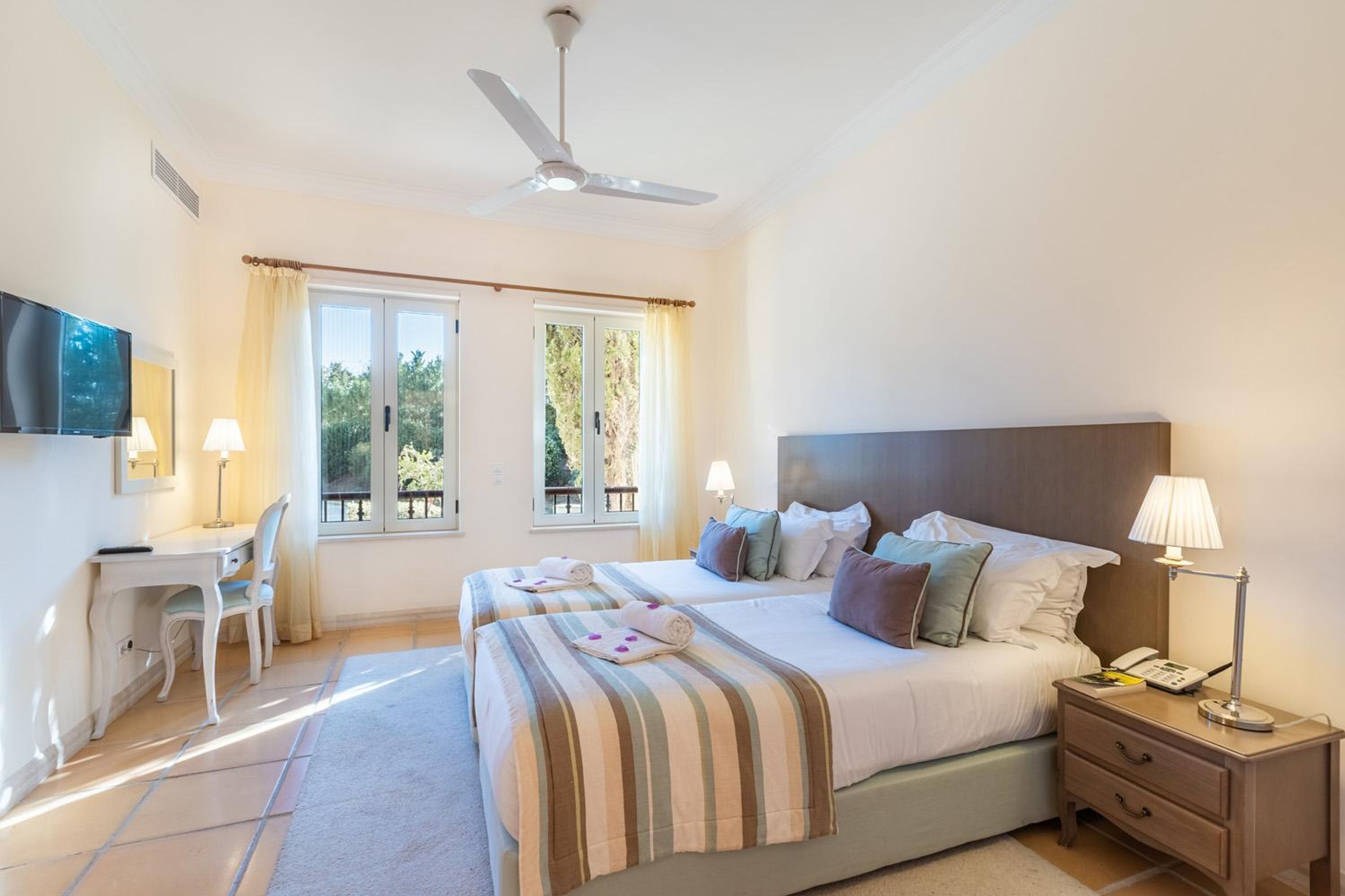 A bedroom with two beds, a desk, a chair, a TV, and a ceiling fan.