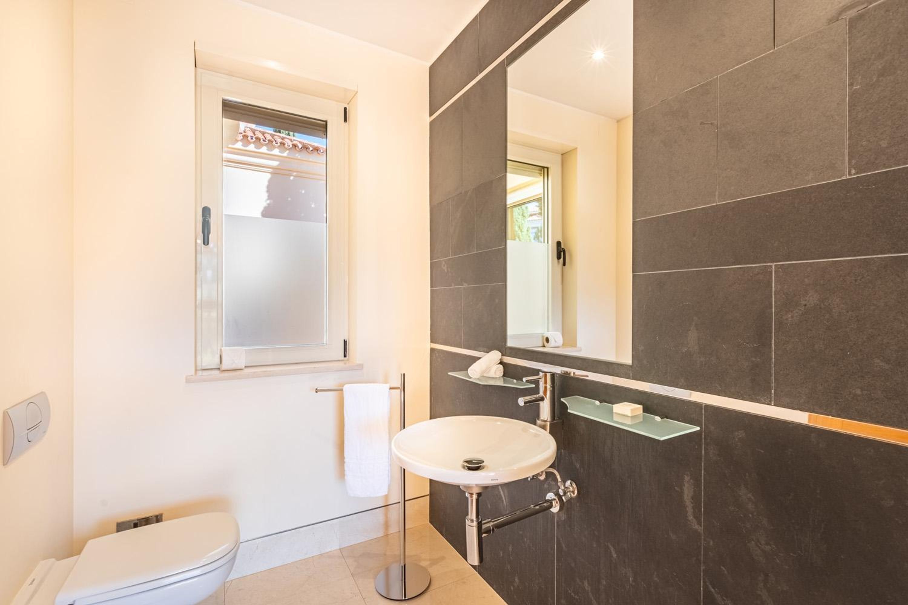 A modern bathroom with a white toilet, a round sink, a large mirror, and dark tiled walls.