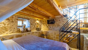 A rustic stone-walled room with a wooden ceiling, a bed, a chair, a small table, a television, and a staircase. - Villa Liana in Spartia (Messini)