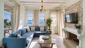 A living room with a blue sofa, a glass coffee table, a TV mounted on a stone wall, and a view of the sea through large windows. - Villa Votsalo in Lefkada (Lefkada)