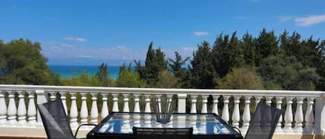 A balcony with a glass table and chairs, overlooking a scenic view of the sea and trees.
