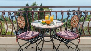 Et rundt bord med to stole, et glas appelsinjuice og en skål med frugt på en balkon med en smuk udsigt.