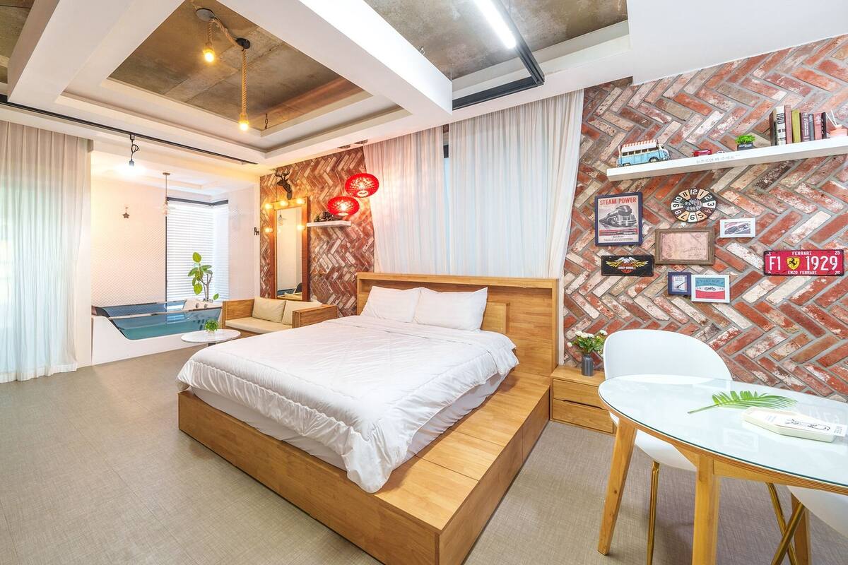 A modern bedroom with a wooden bed, a small seating area, a glass table, and a brick-patterned wall with various decorations.