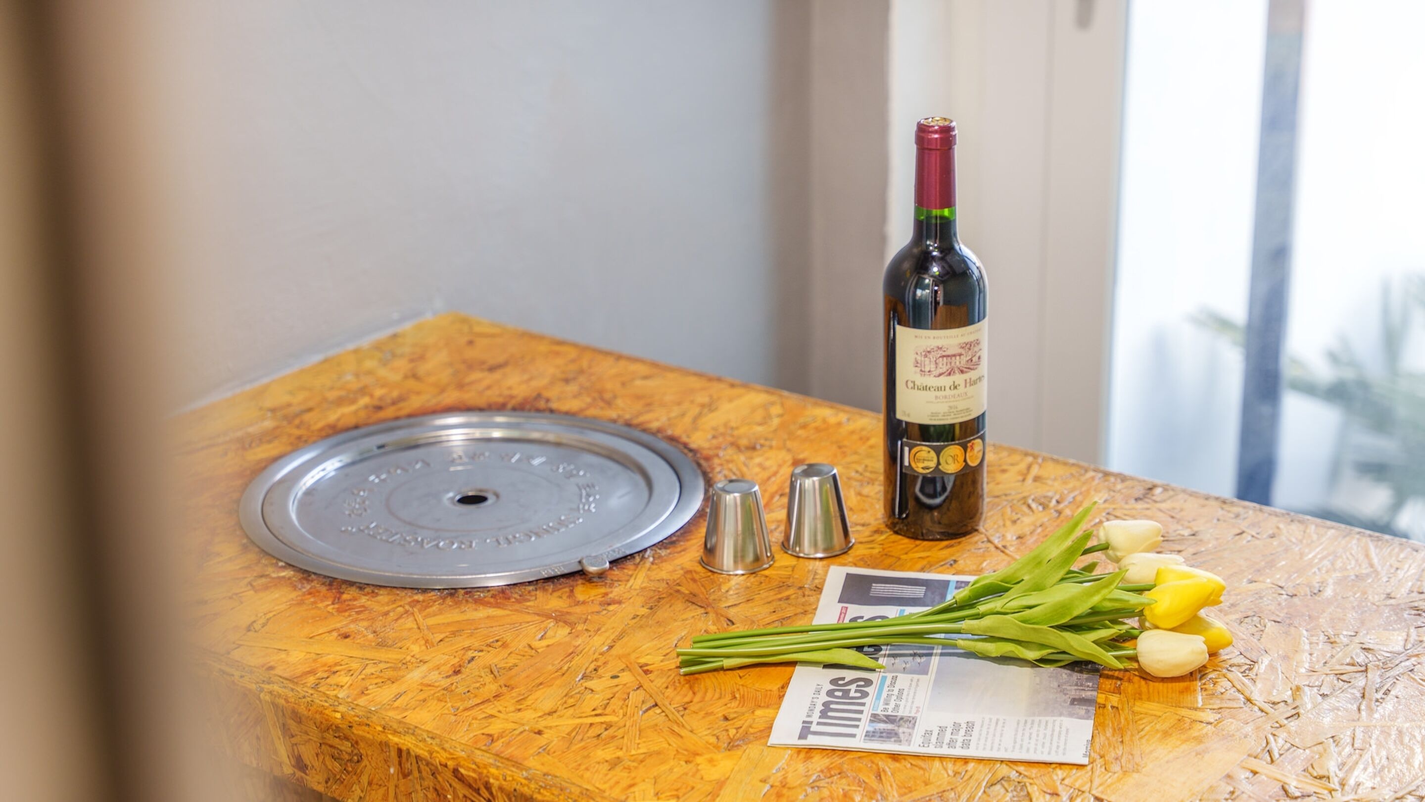 Una mesa de madera con una botella de vino, saleros y pimenteros, un periódico y un ramo de flores.