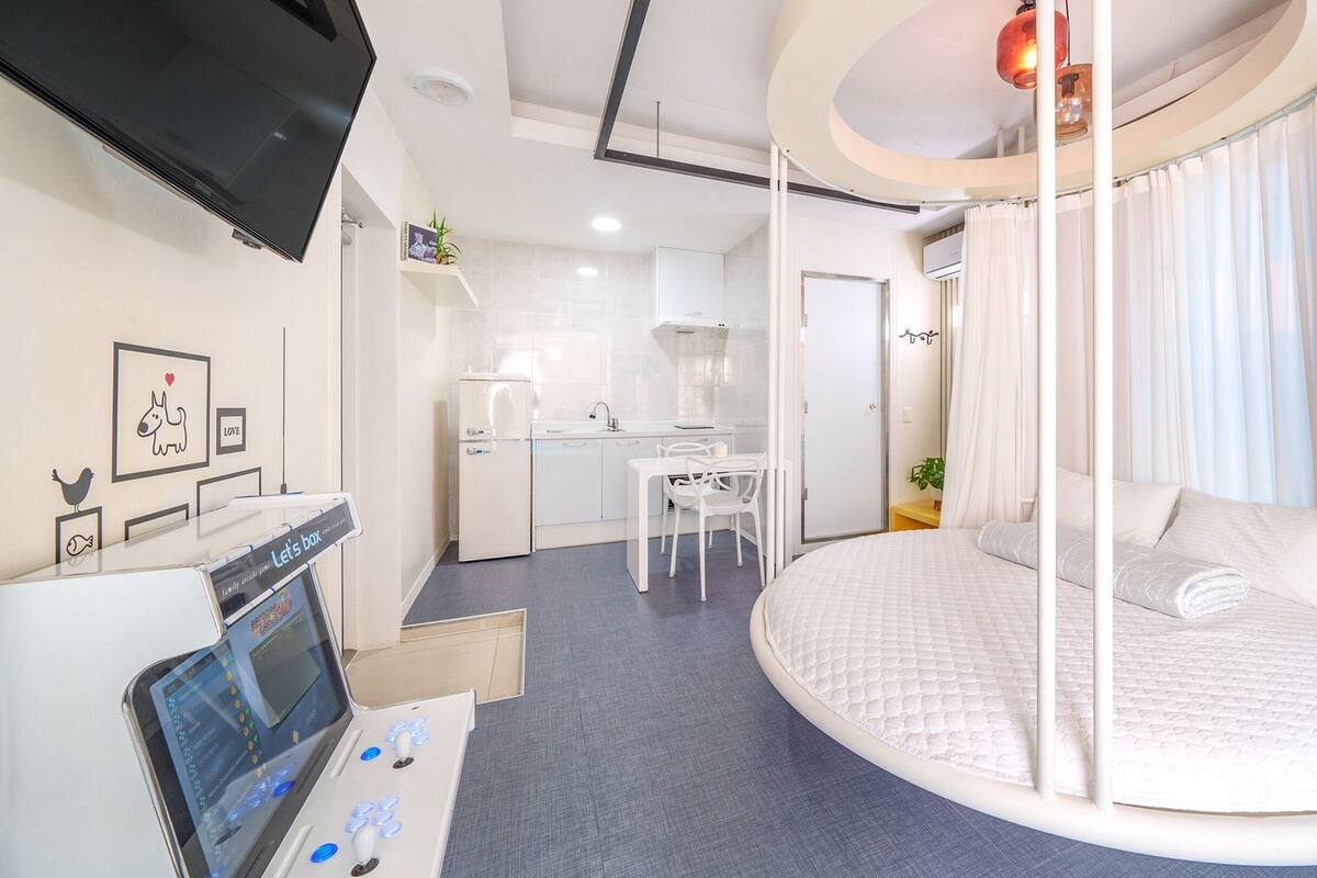 A modern hotel room with a circular bed, a small kitchenette, a desk, and a TV.