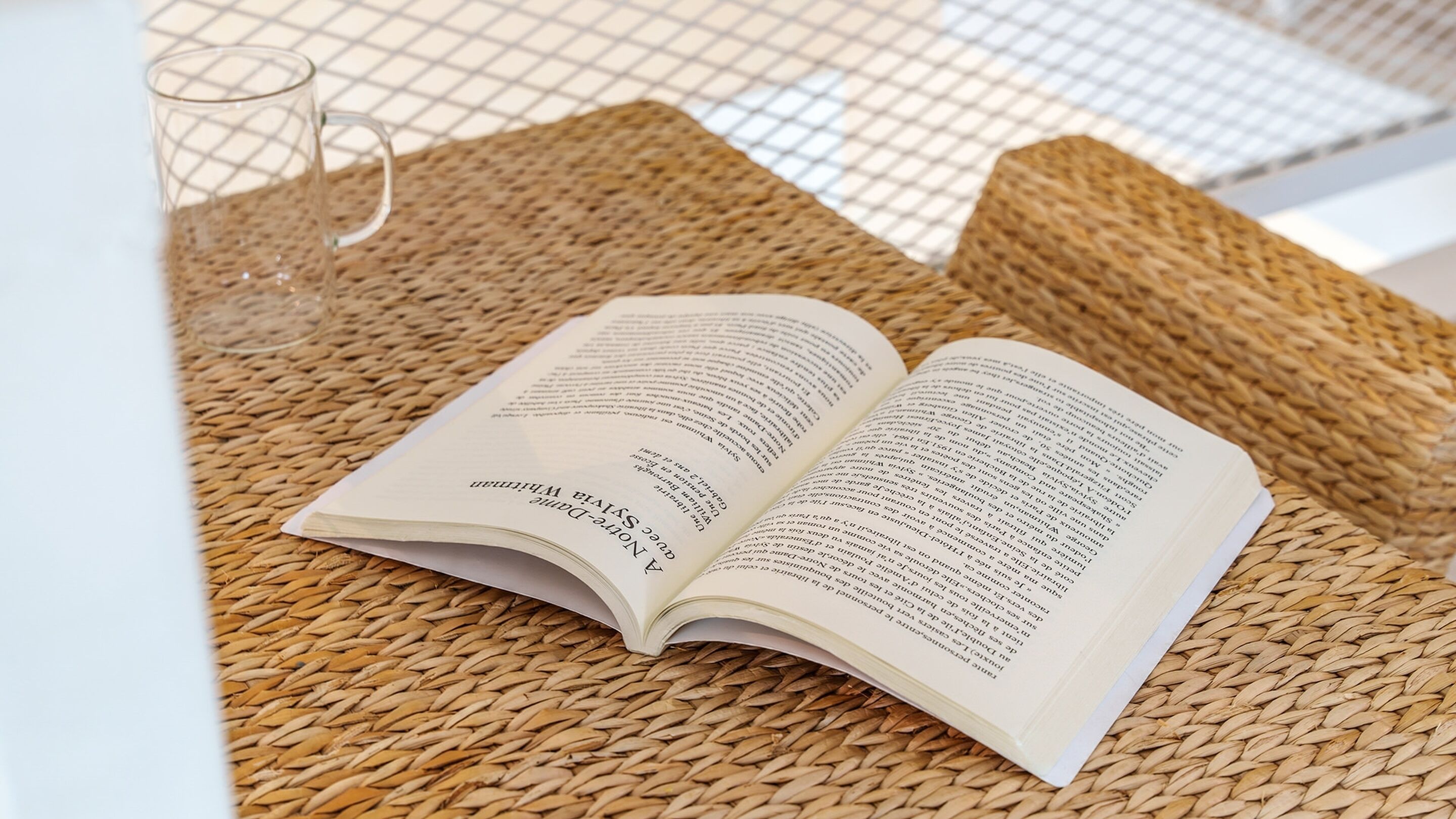 An open book on a woven surface with a clear glass beside it.