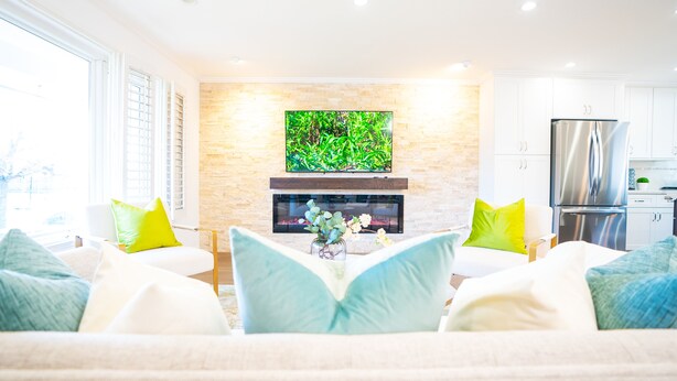 A modern living room with a fireplace, a flat-screen TV mounted above it, and a kitchen area to the right.