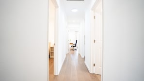 A bright hallway with white walls and wooden flooring, leading to a room with a desk and chair.