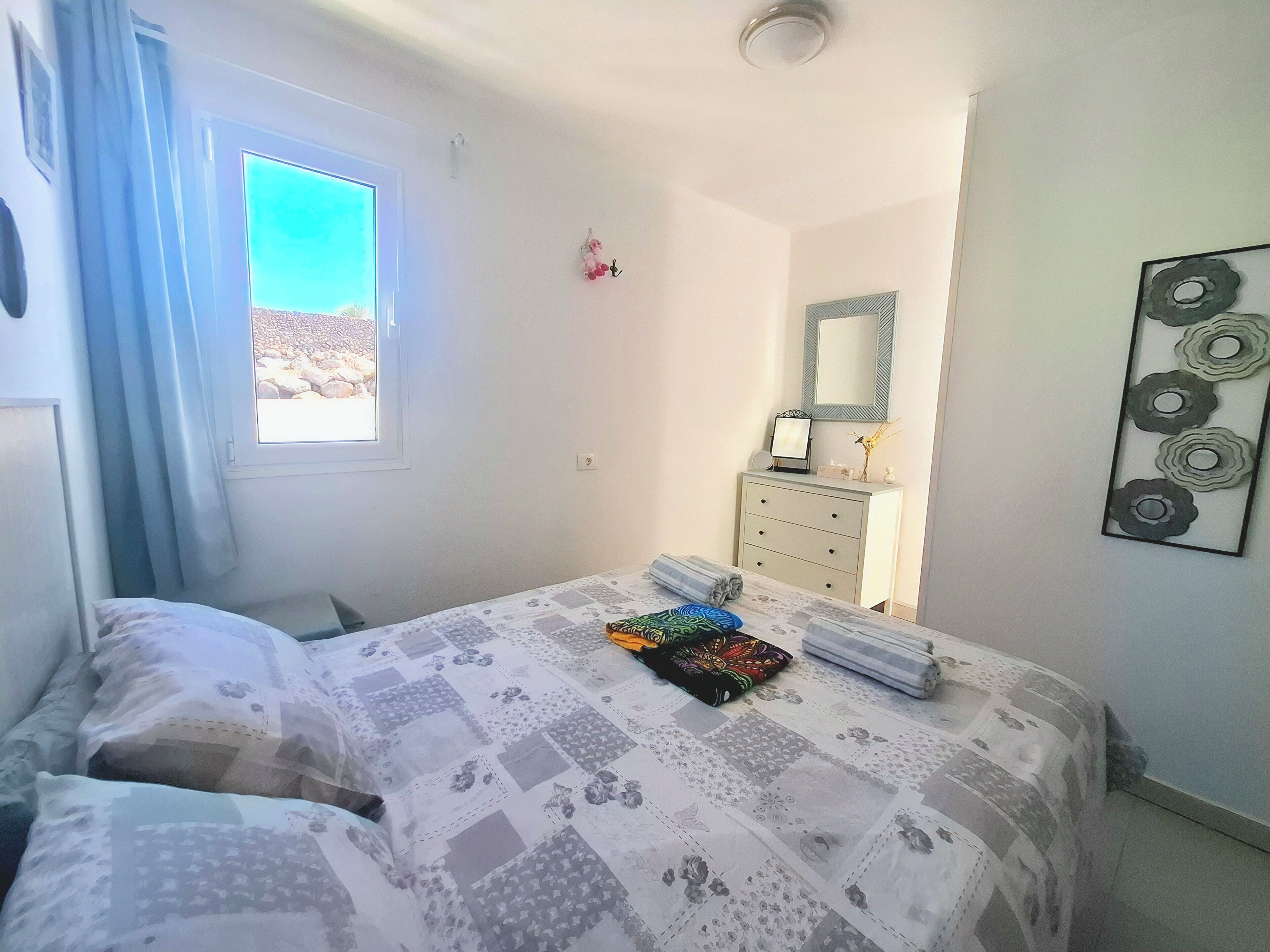 Une chambre à coucher comprenant un lit, une commode, une fenêtre avec des rideaux, un miroir et une œuvre d’art murale.