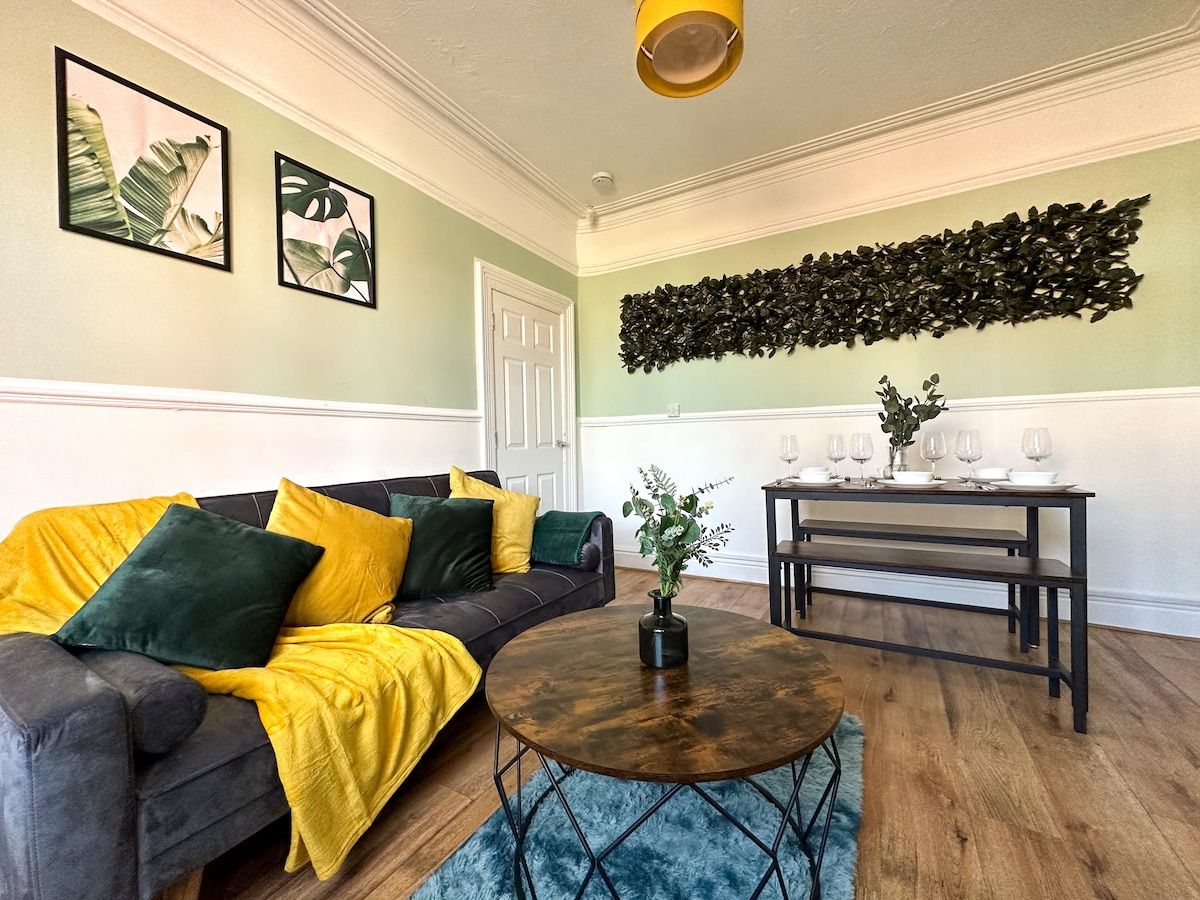 A living room with a sectional sofa, a round coffee table, a wall-mounted plant, and a bench with wine glasses.