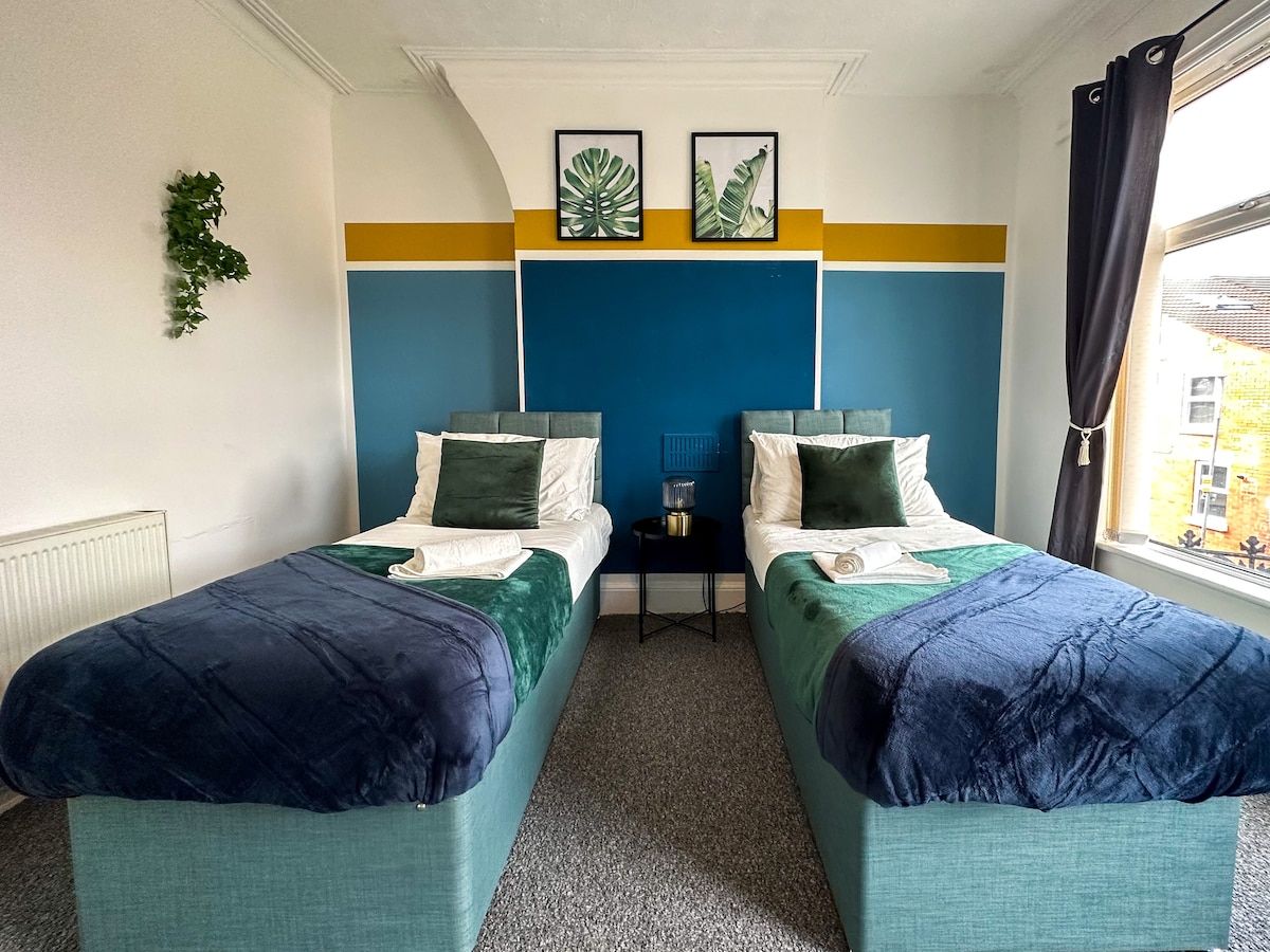 A twin room with two single beds, a small table, a plant, and a window with curtains.