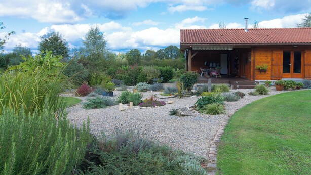 A wooden cabin with a red-tiled roof, surrounded by a well-maintained garden with various plants and a gravel pathway. - Country cottage (Linards)