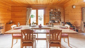 A wooden cabin with a dining table set for four, surrounded by chairs, and a bookshelf filled with books and decorative items. - Country cottage (Linards)