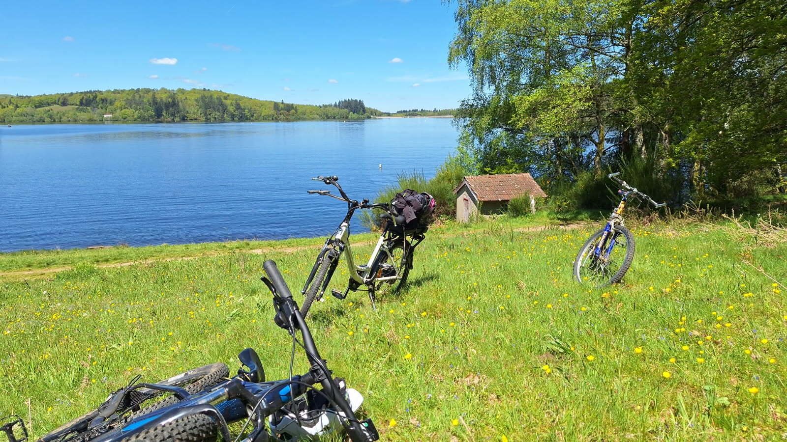 Blick auf einen See mit Fahrrädern, die auf der Wiese stehen, eine kleine Werkstatt und einem strahlend blauen Himmel.