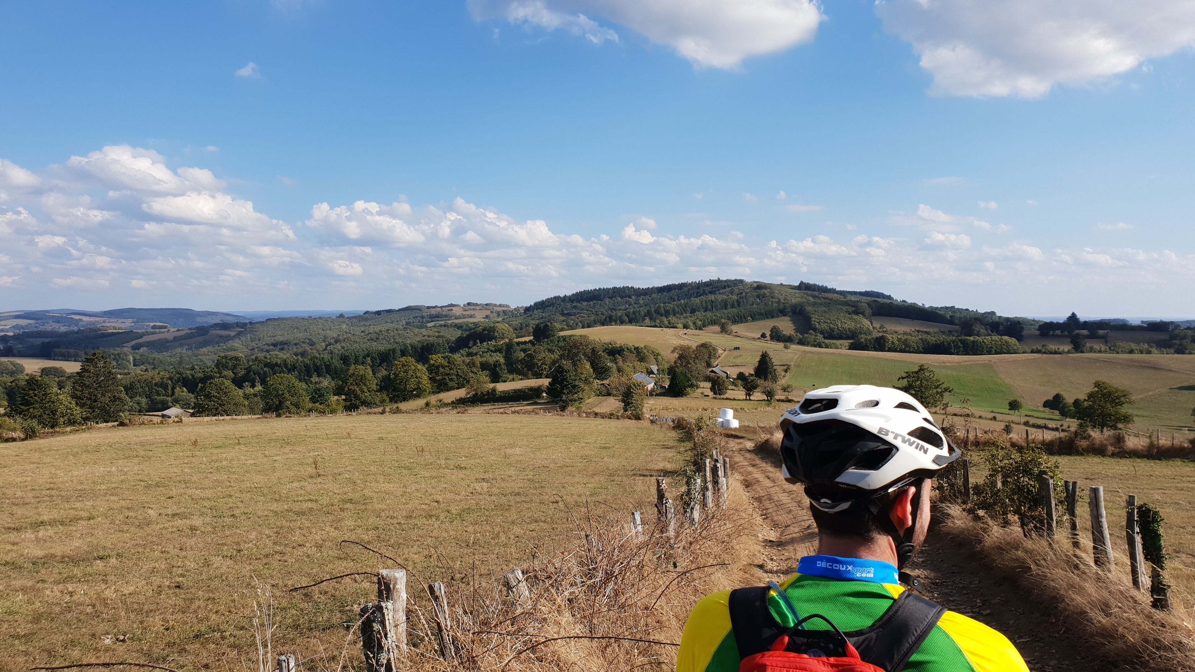 Un cycliste sur un sentier de campagne, avec en toile de fond des collines ondoyantes et quelques maisons dispersées.