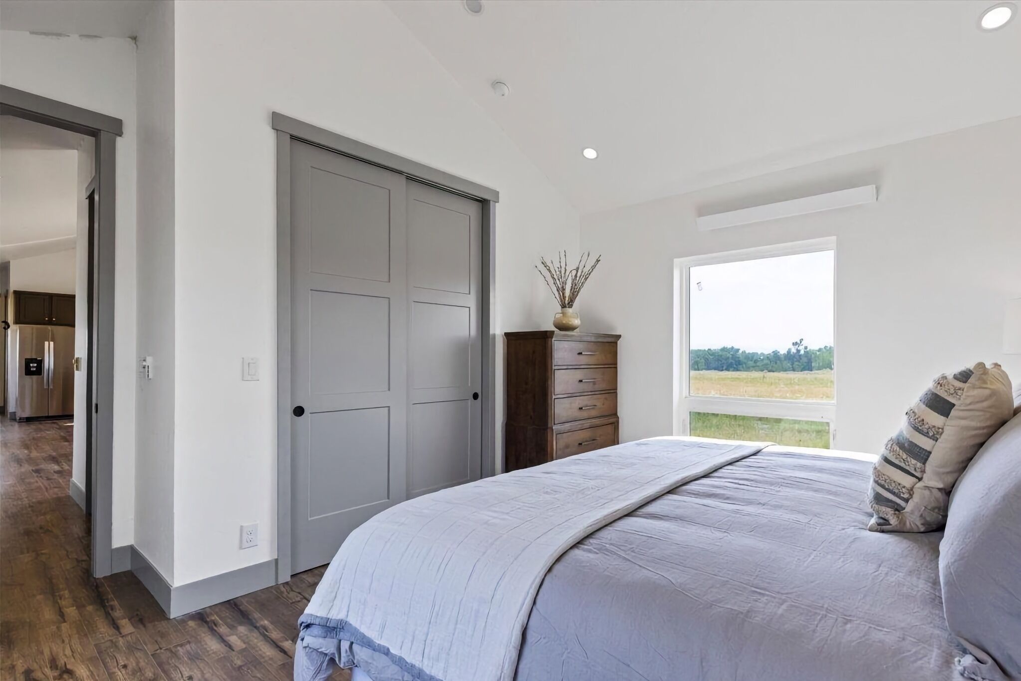 A bedroom with a large bed, a dresser, a window with a view of the outdoors, and a door leading to another room.