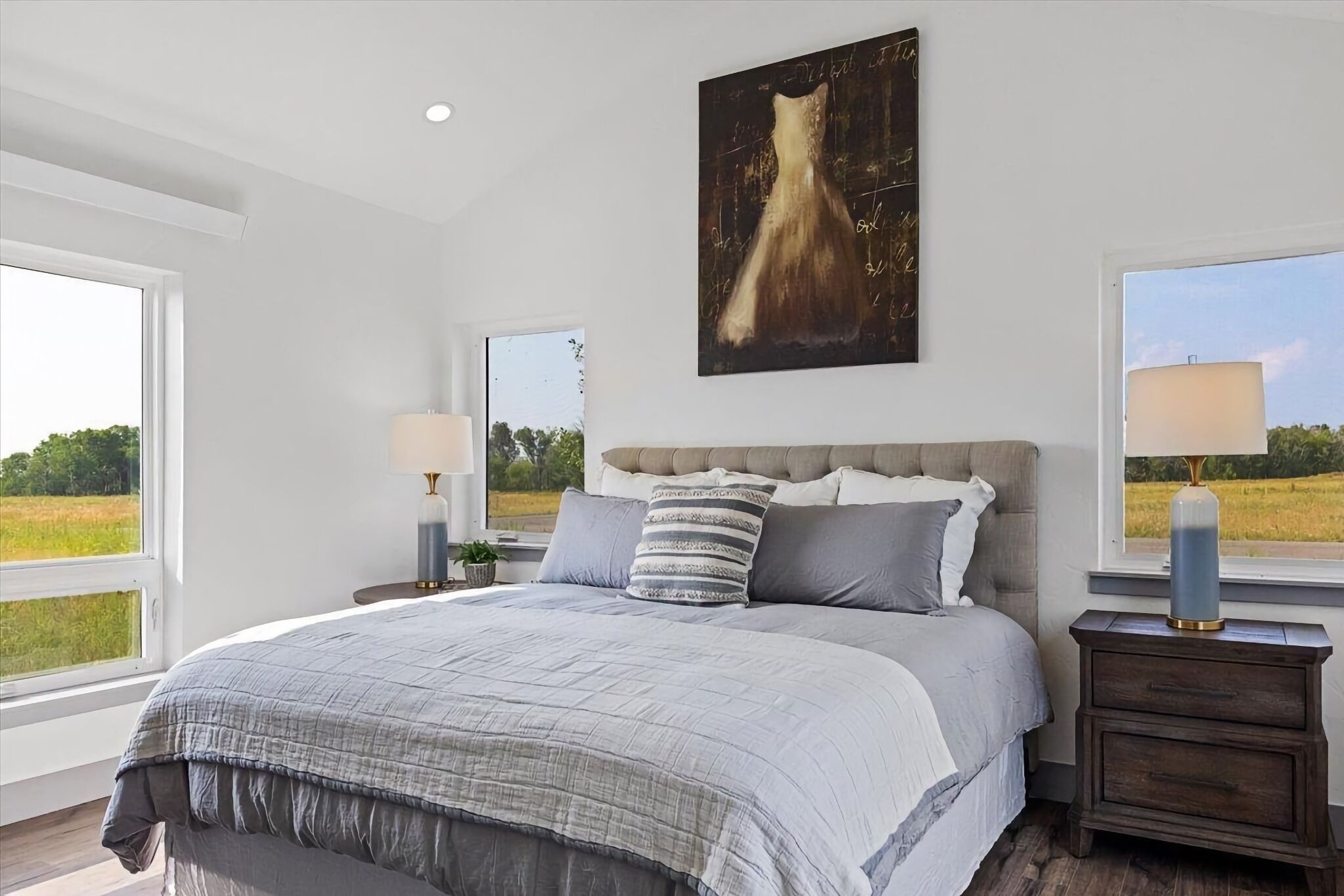 A bedroom with a large bed, a headboard, two bedside tables, a nightstand, a window, and a painting on the wall.