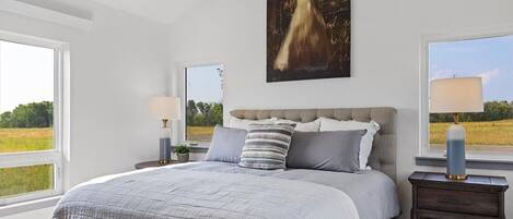 A bedroom with a large bed, a headboard, two bedside tables, a nightstand, a window, and a painting on the wall.