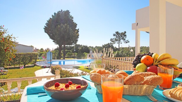 A breakfast table with fruits, pastries, and orange juice on a balcony overlooking a swimming pool and garden. - Villa Bella Vista (Albufeira)