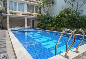 Une piscine rectangulaire aux carreaux bleus et dotée de rampes métalliques, entourée d’une terrasse en béton et bordée d’un bâtiment avec des balcons.